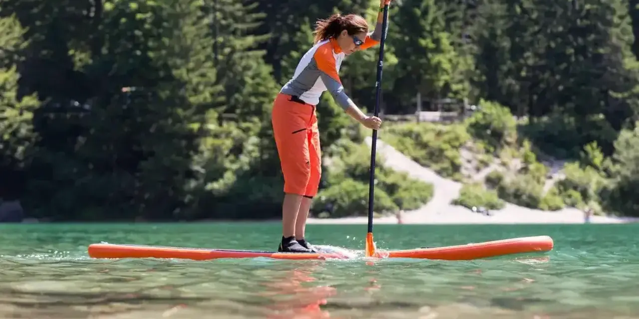 Kobieta w pomarańczowych spodenkach i sportowej bluzce uprawia pływanie na supie po spokojnym jeziorze. W tle zielone drzewa i jasna plaża.