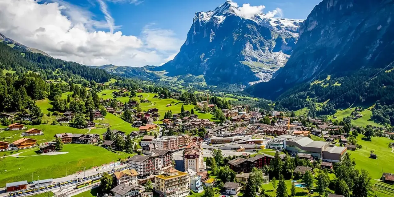 A scenic view of Grindelwald, Switzerland, nestled in a valley with lush green hills and a majestic snow-capped mountain. A train travels through the village.