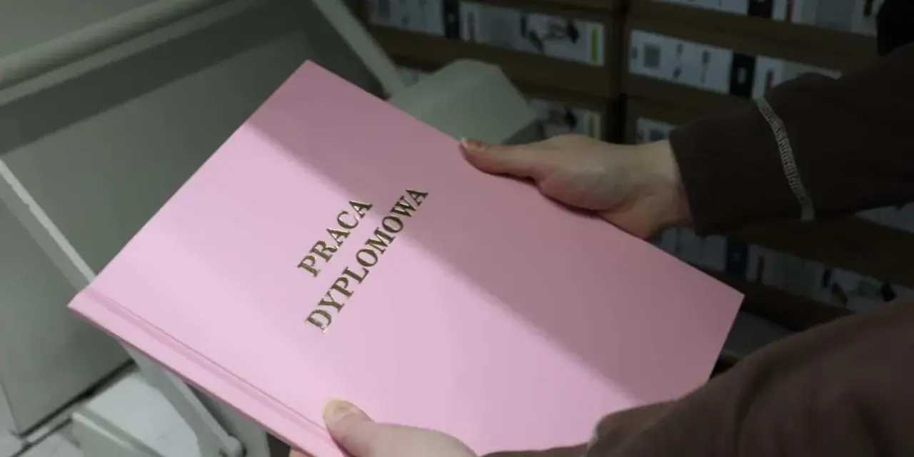 Różowa okładka z napisem "PRACA DYPLOMOWA" w złotych literach, trzymana w dłoniach nad bindownicą.
