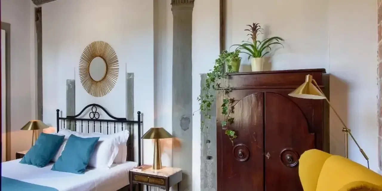 Elegancka sypialnia z łóżkiem, zabytkową szafą i żółtym fotelem. Idealne miejsce, by dowiedzieć się, gdzie nocować w Toskanii.