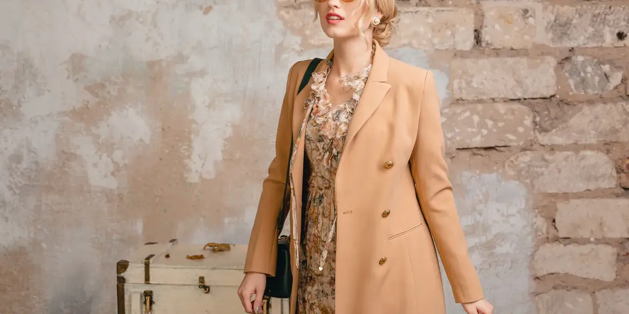 Elegancka kobieta w płaszczu camelowy kolor, sukience w kwiaty i okularach przeciwsłonecznych pozuje przy starej walizce.
