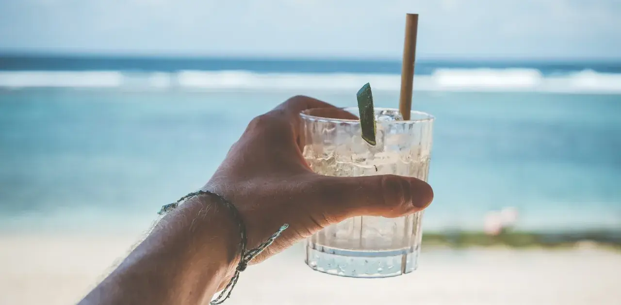 Orzeźwiający drink na plaży, z lodem i plasterkiem limonki, w dłoni z bransoletką. W tle błękitne morze i fale.