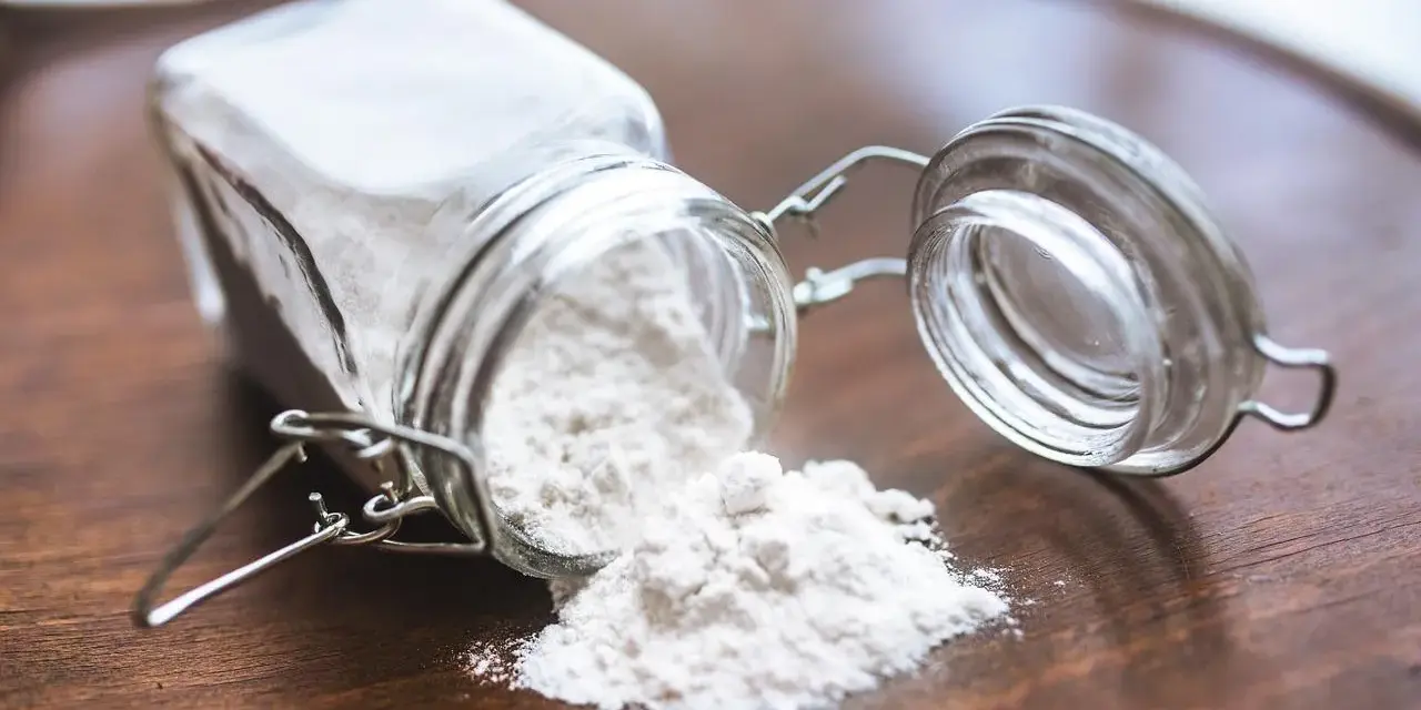 Un barattolo di vetro rovesciato su un tavolo di legno, con polvere bianca che fuoriesce, forse ingredienti per colla vinilica fatta in casa.