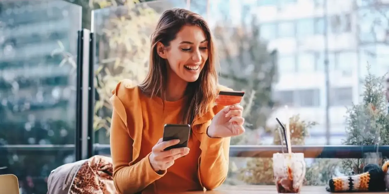 Smiling woman uses phone and credit card. She's researching if you need an LLC for a business credit card.