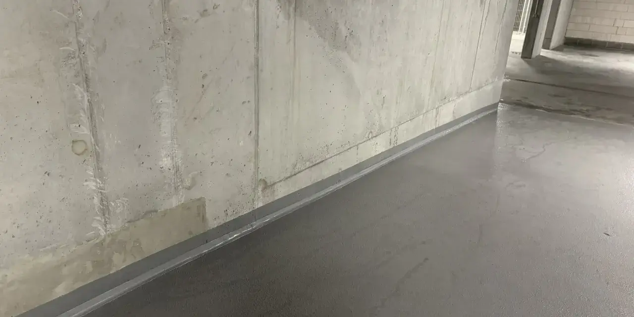 Szara, gładka posadzka w garażu, zabezpieczająca co na podłogę w garażu przed wilgocią. Betonowa ściana w tle.