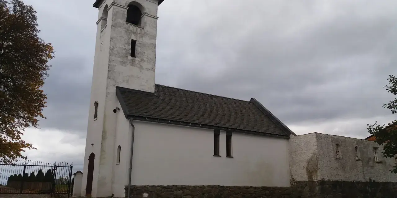 Biały, zabytkowy dwór z wieżą i dachem krytym dachówką. W oddali widać bramę i drzewa.