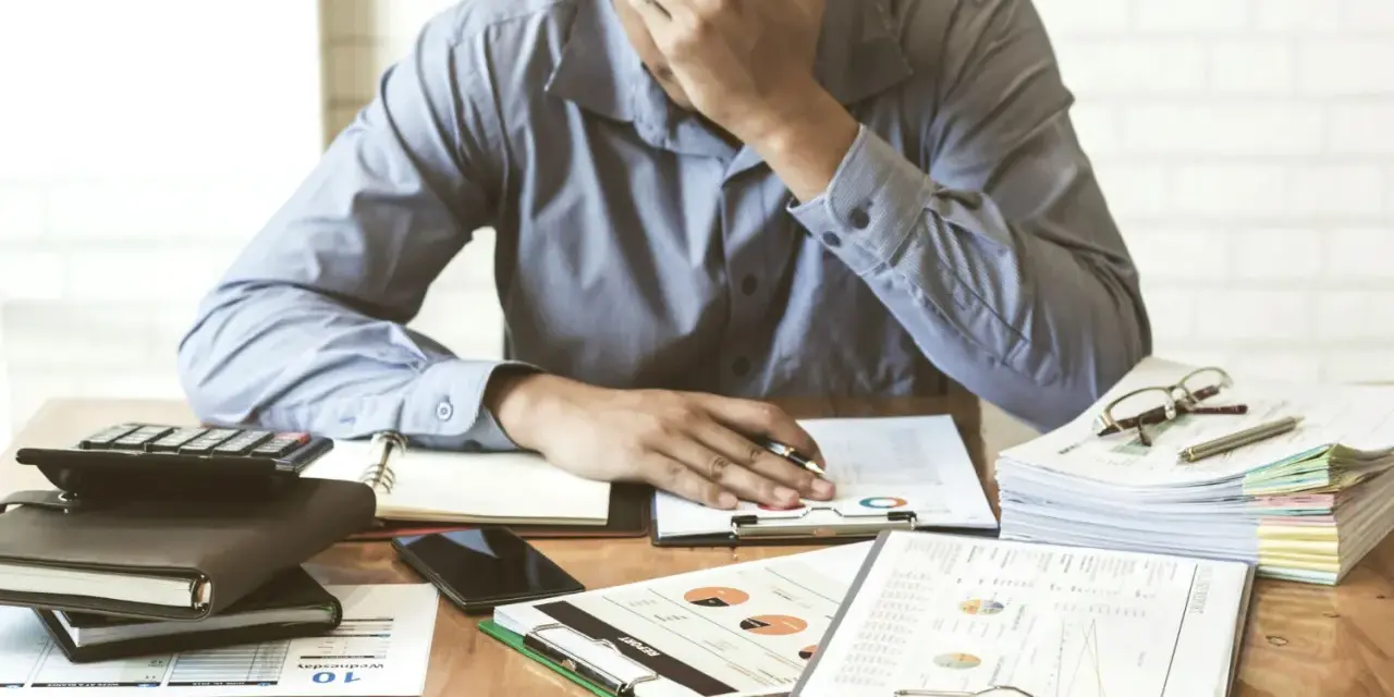 Zestaw dokumentów, kalkulator i telefon leżą na biurku. Mężczyzna z ręką na czole, przytłoczony problemami finansowymi, przegląda pismo windykacyjne.
