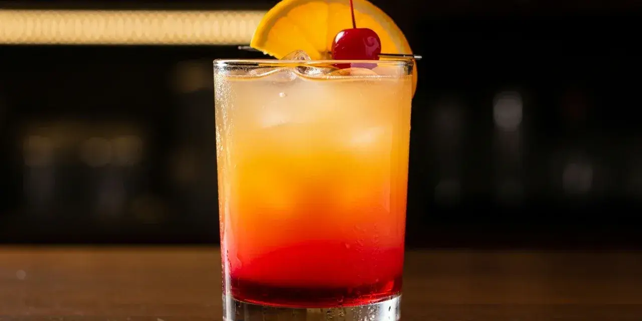 En klassisk tequila sunrise med lager av rött och orange, garnerad med apelsinskiva och körsbär.