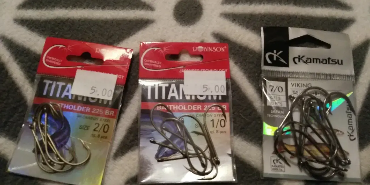 Trzy opakowania haczyków wędkarskich: dwa z napisem "TITANIUM" i jeden z logo "Kamatsu".