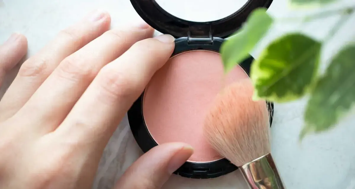 Une main applique un pinceau sur un blush poudre, révélant une douce teinte couleur blush.