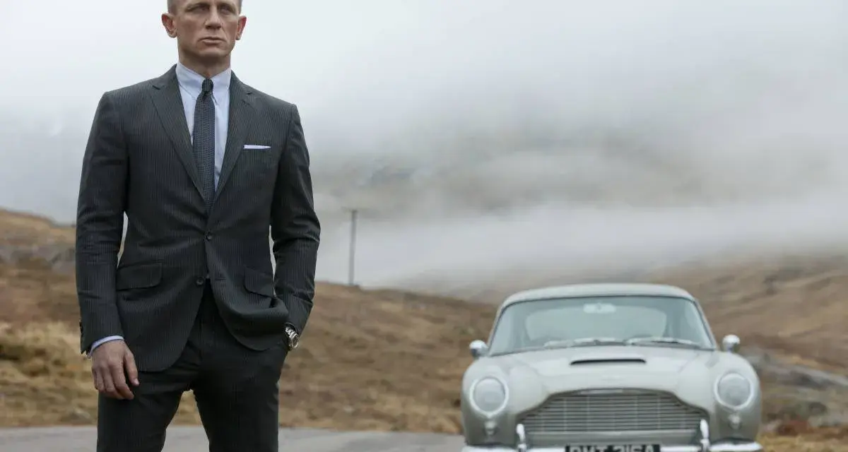 Uomo con impeccabile 007 outfit, completo gessato e cravatta, posa accanto a una Aston Martin d'epoca in un paesaggio nebbioso.