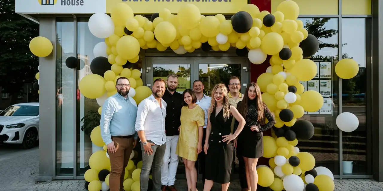 Grupa ludzi pozuje przed biurem nieruchomości ozdobionym żółtymi balonami.