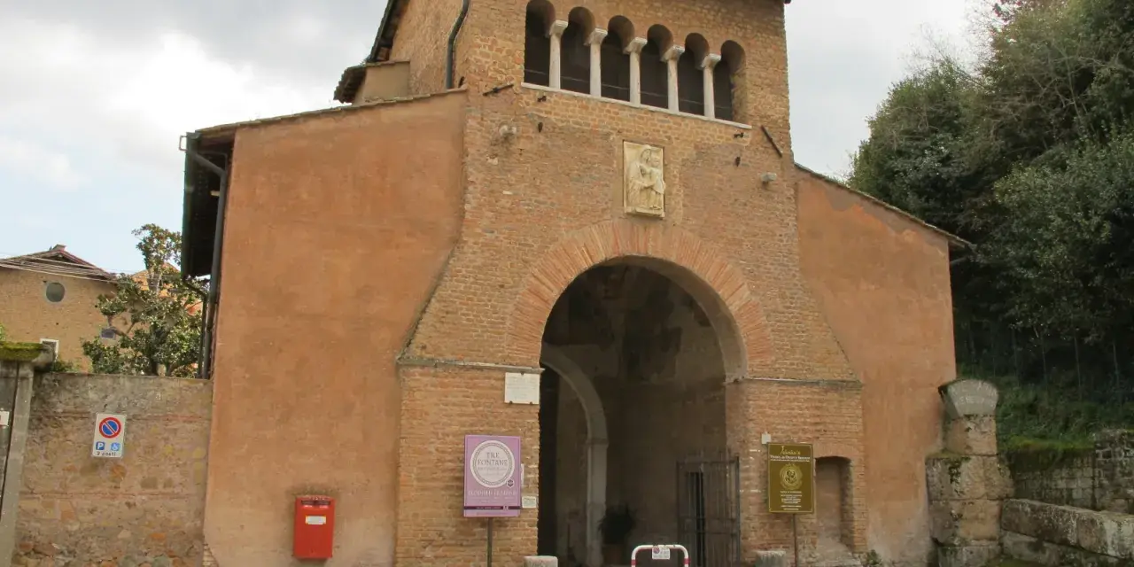 The imposing facade of Tre Fontane Abbey, with its arched entrance and upper loggia, stands against a cloudy sky.