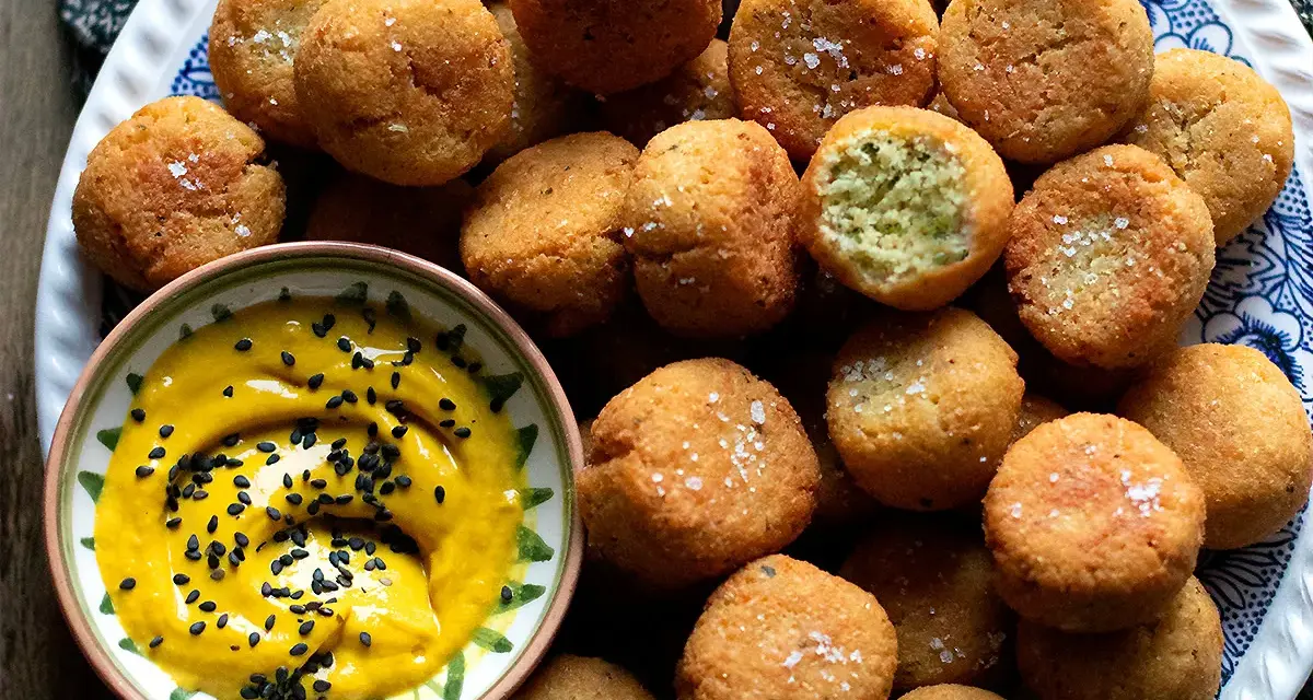 Falafel ricetta originale: croccanti palline dorate, una con un morso rivela il verde interno, accompagnate da una salsa gialla con semi di sesamo nero.