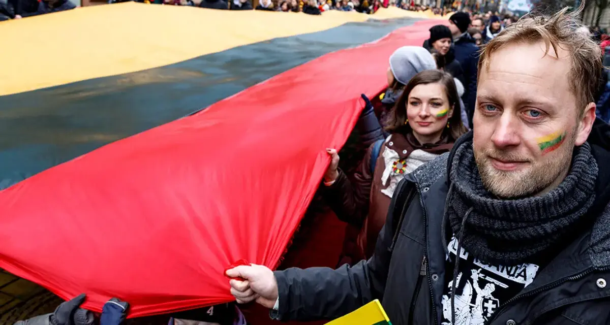 Ludzie niosą ogromną flagę Litwy podczas święta narodowego. Na twarzach uczestników widać patriotyczne barwy.