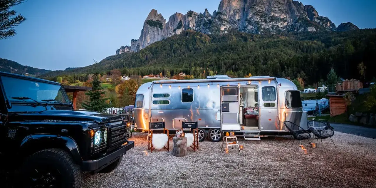 Ein Airstream-Wohnwagen und ein schwarzer Geländewagen stehen auf einem **Campingplatz Dolomiten** mit majestätischen Bergen im Hintergrund. Gemütliche Stühle und Lichter schaffen eine einladende Atmosphäre.