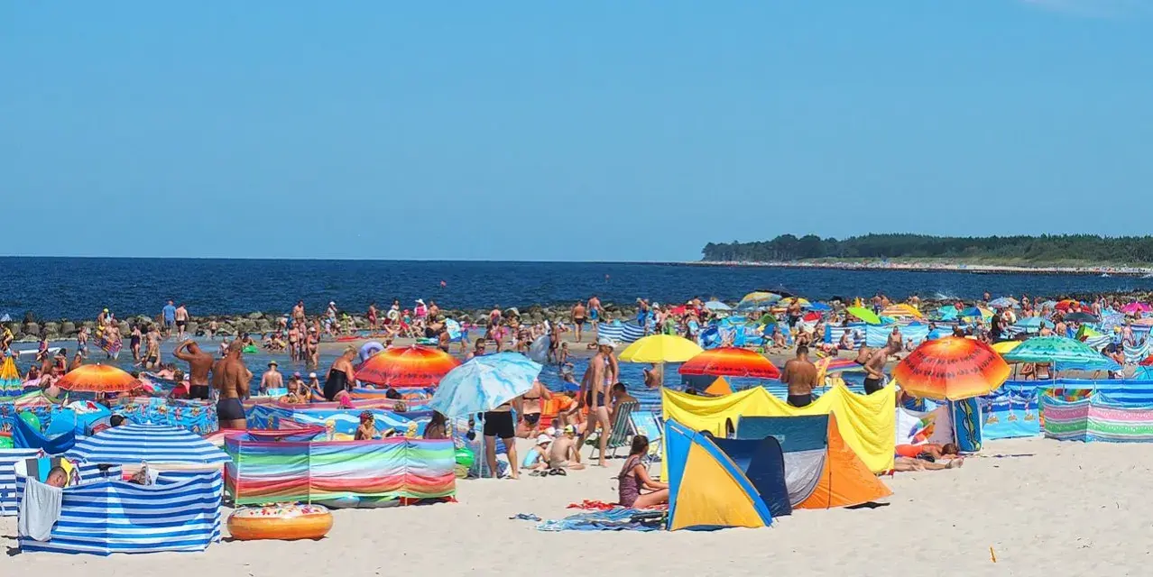 Tłum ludzi na plaży w Darłówku Wschodnim, kolorowe parawany i namioty chronią przed słońcem.