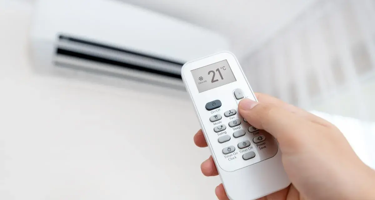 Mano ajustando el aire acondicionado a 21°C en modo "Cooling". La pantalla del control remoto muestra la temperatura y el modo, indicando que significa "cool" en el aire acondicionado: enfriar.