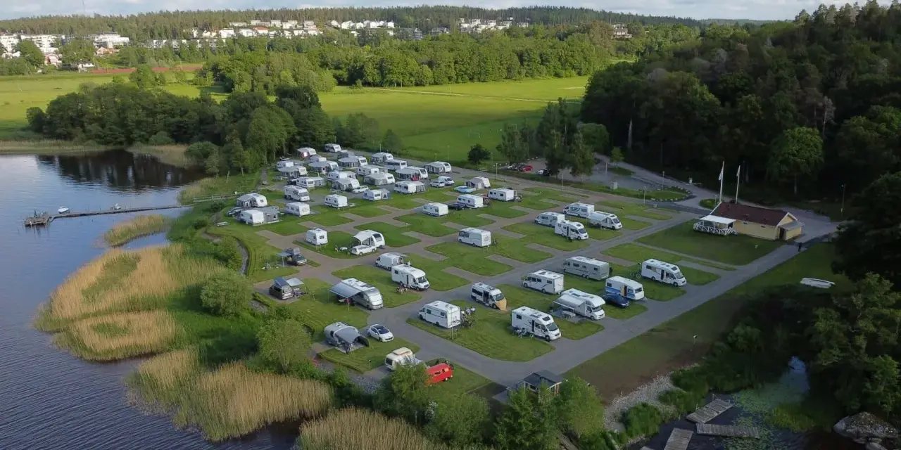 Full campingplats med husbilar och husvagnar vid en sjö, med grönskande träd och bebyggelse i bakgrunden. En idyllisk plats för camping i Alingsås.