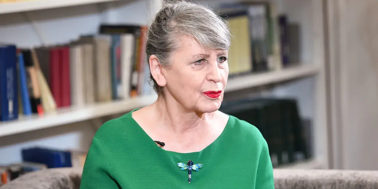 Joanna Wnuk-Nazarowa w zielonej bluzce z broszką w kształcie ważki, na tle regałów z książkami.