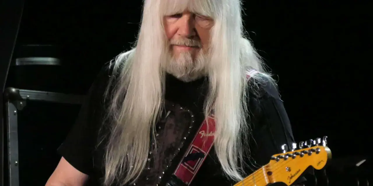 Andy Scott mit seinen charakteristischen langen, weißen Haaren spielt auf einer orangen E-Gitarre.