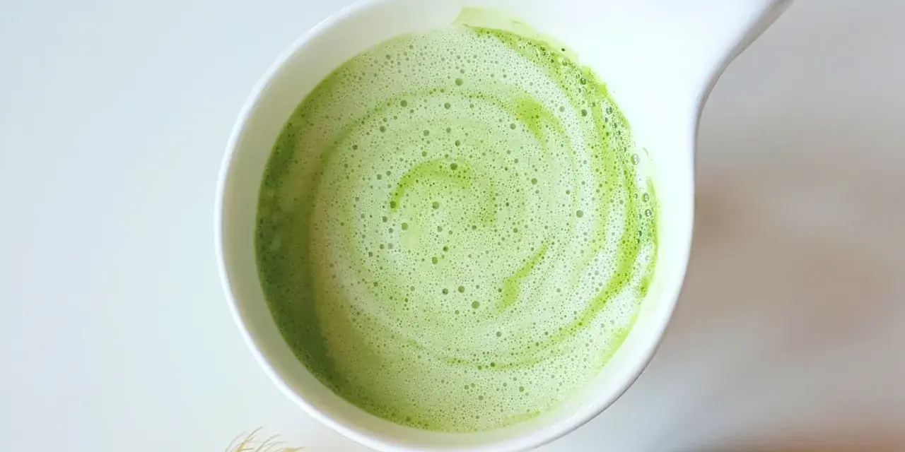 Zielony, spieniony napój matcha w białej miseczce, obok bambusowa miotełka i puszka z proszkiem. Idealne co na pobudzenie zamiast kawy.