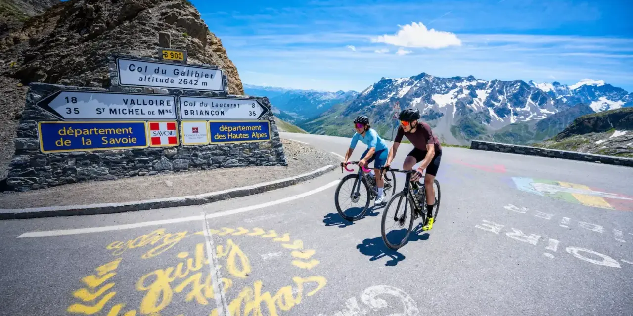 Ciclisti in salita verso il Col du Galibier, con montagne innevate sullo sfondo e segnaletica stradale.