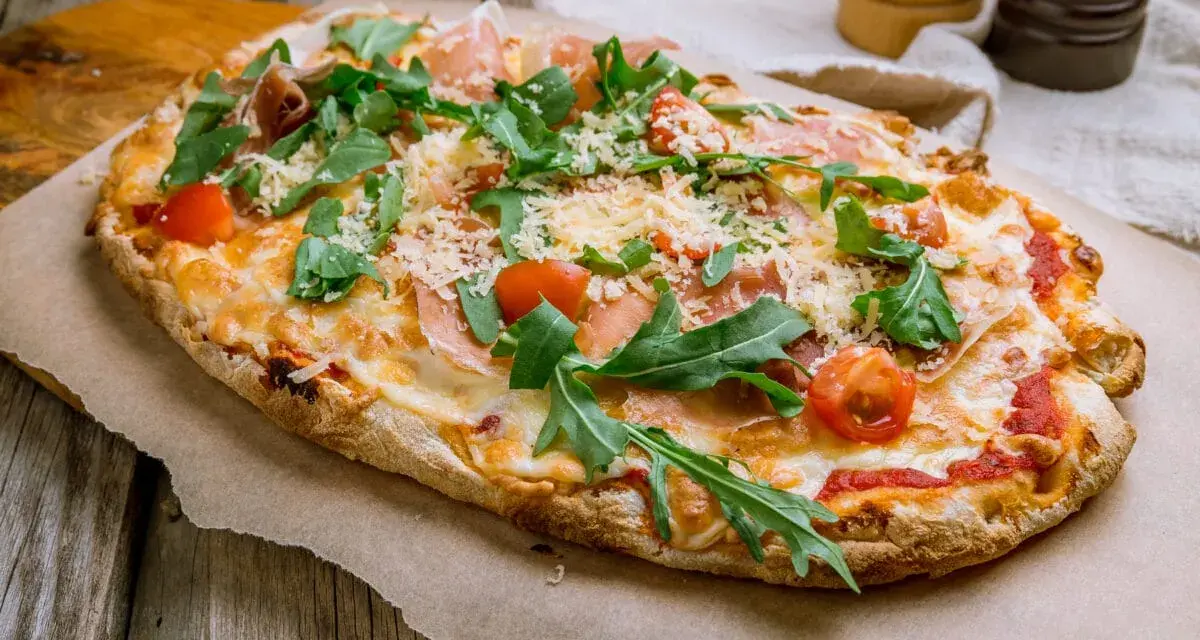 Pinsa co to? Aromatyczna, podłużna pizza z rukolą, pomidorkami i szynką parmeńską, posypana parmezanem.