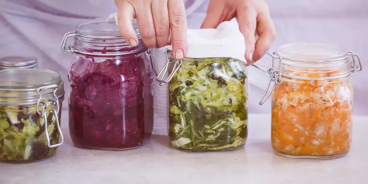 Hände bedecken ein Glas mit fermentiertem Gemüse, Teil einer Auswahl an probiotischen Lebensmitteln in Gläsern.