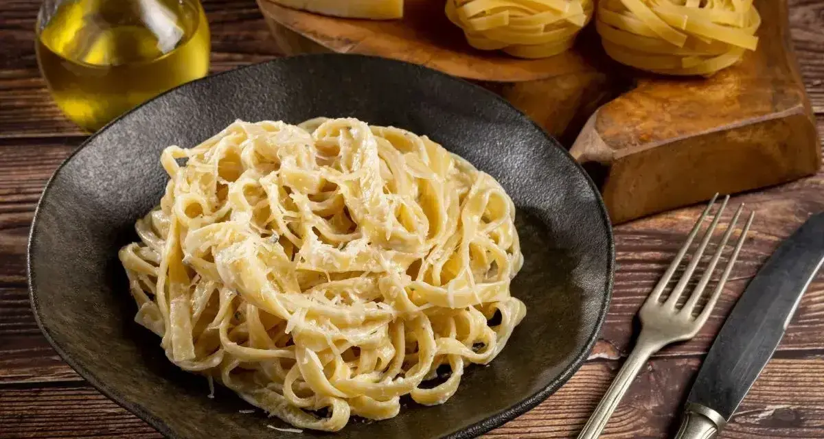 Pyszne Fettuccine Alfredo przepis na talerzu, obok surowy makaron, ser i oliwa. Idealne danie dla miłośników włoskiej kuchni.