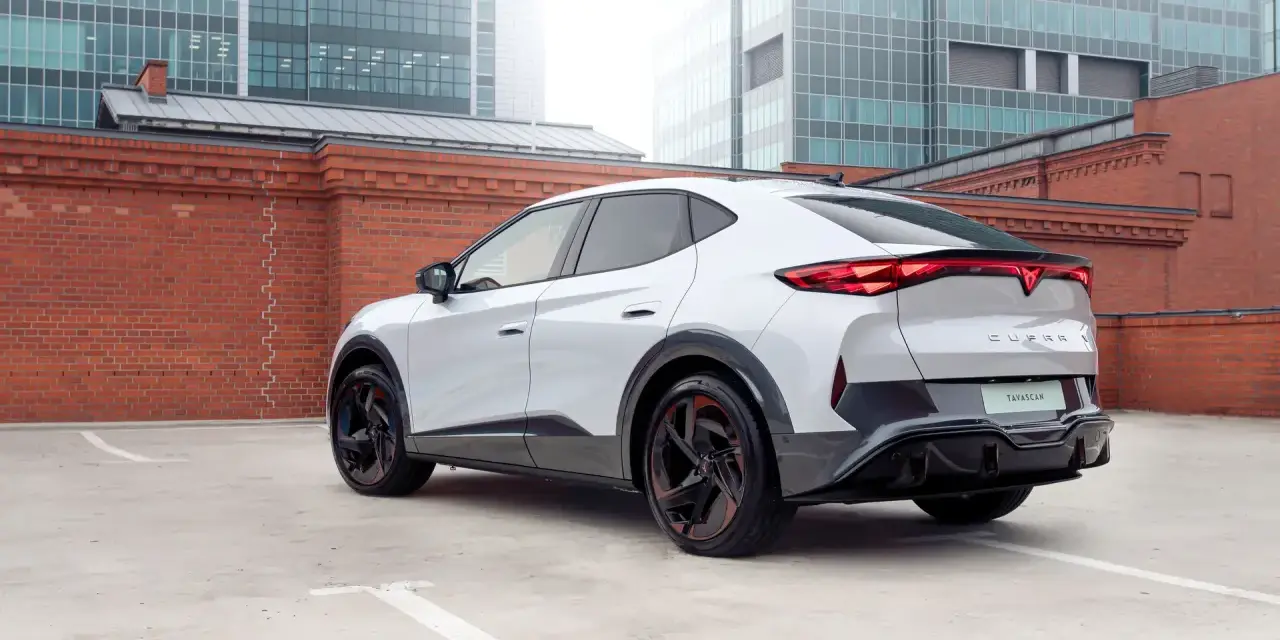 Biały elektryczny SUV Cupra Tavascan z czerwonymi akcentami na felgach, zaparkowany na tle ceglanego muru i nowoczesnych budynków.
