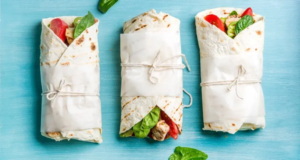 Trzy keto tortille z siemienia lnianego, zawinięte w papier i przewiązane sznurkiem, z widocznym nadzieniem z kurczaka, pomidorów i szpinaku.