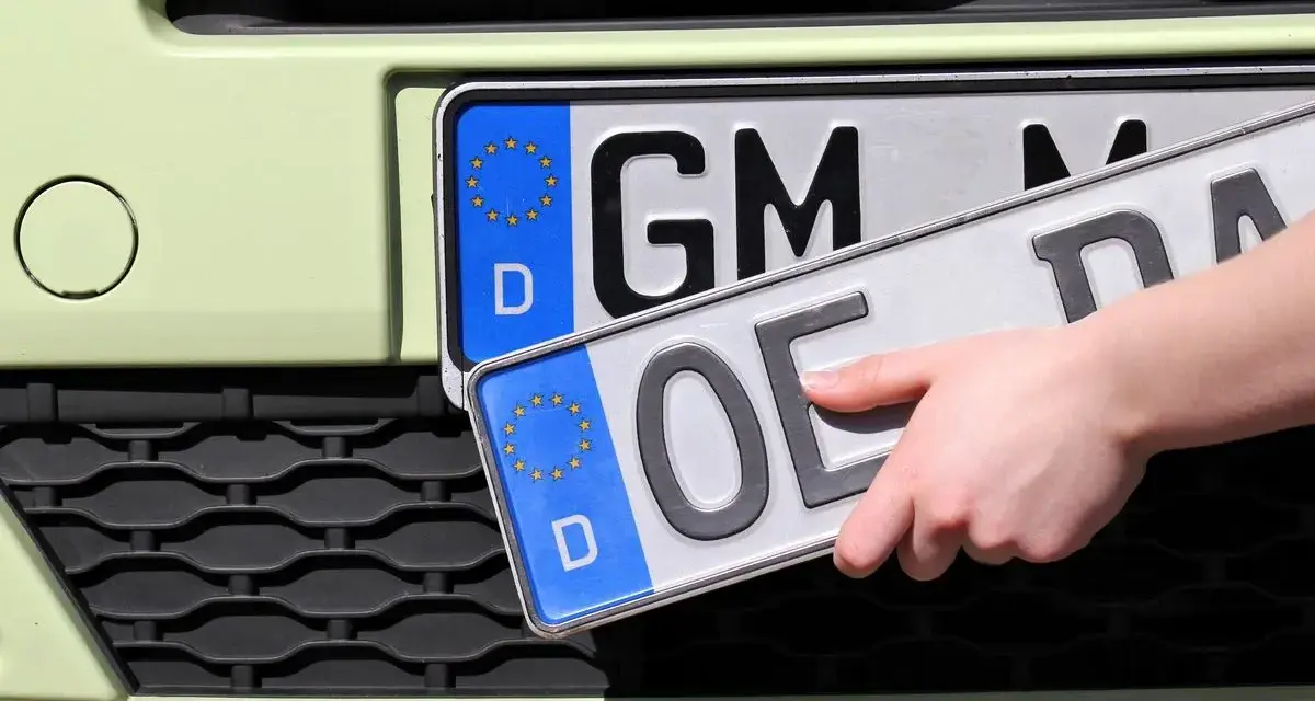 Dłoń trzyma nową tablicę rejestracyjną z literami "OE", gotową do zamontowania na samochodzie.
