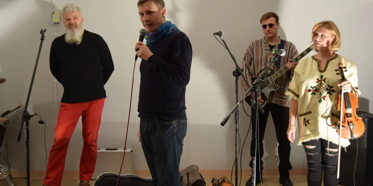 Orkiestra Świętego Mikołaja przygotowuje się do występu. Wokalista z mikrofonem, gitarzysta, skrzypaczka i brodaty mężczyzna w czerwonych spodniach.
