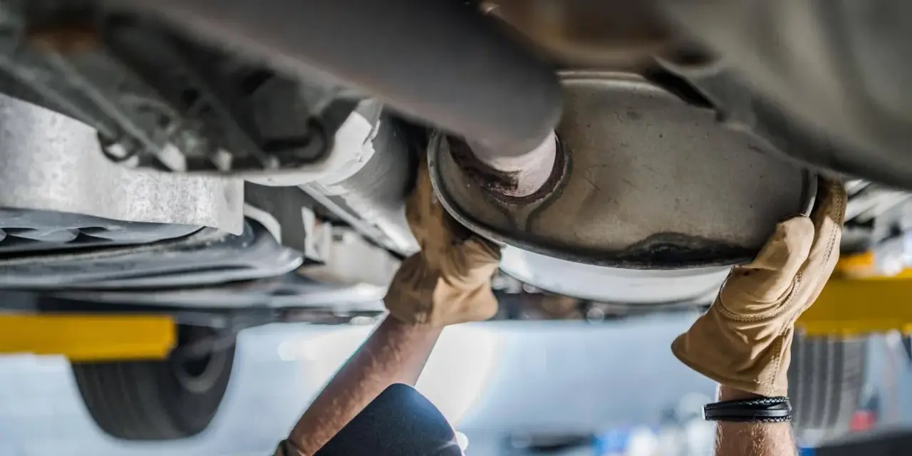 Mani guantate lavorano sul sistema di scarico di un'auto, forse sostituendo il catalizzatore auto.