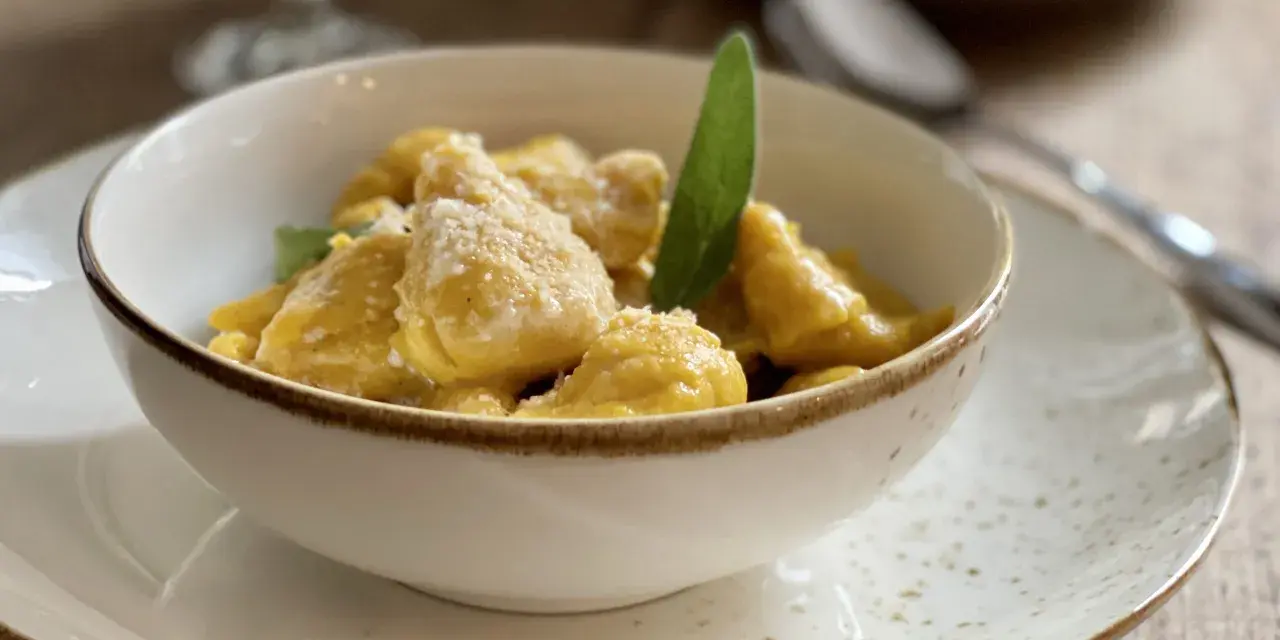 Gnocchi di zucca dorati e soffici, conditi con parmigiano e una foglia di salvia, serviti in una ciotola bianca su un piatto maculato.