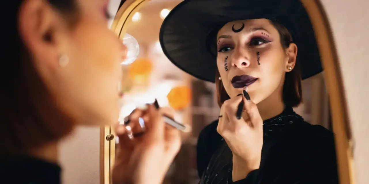 Frau mit Hexenhut und Halloween-Make-up trägt Lippenstift vor einem Spiegel auf.