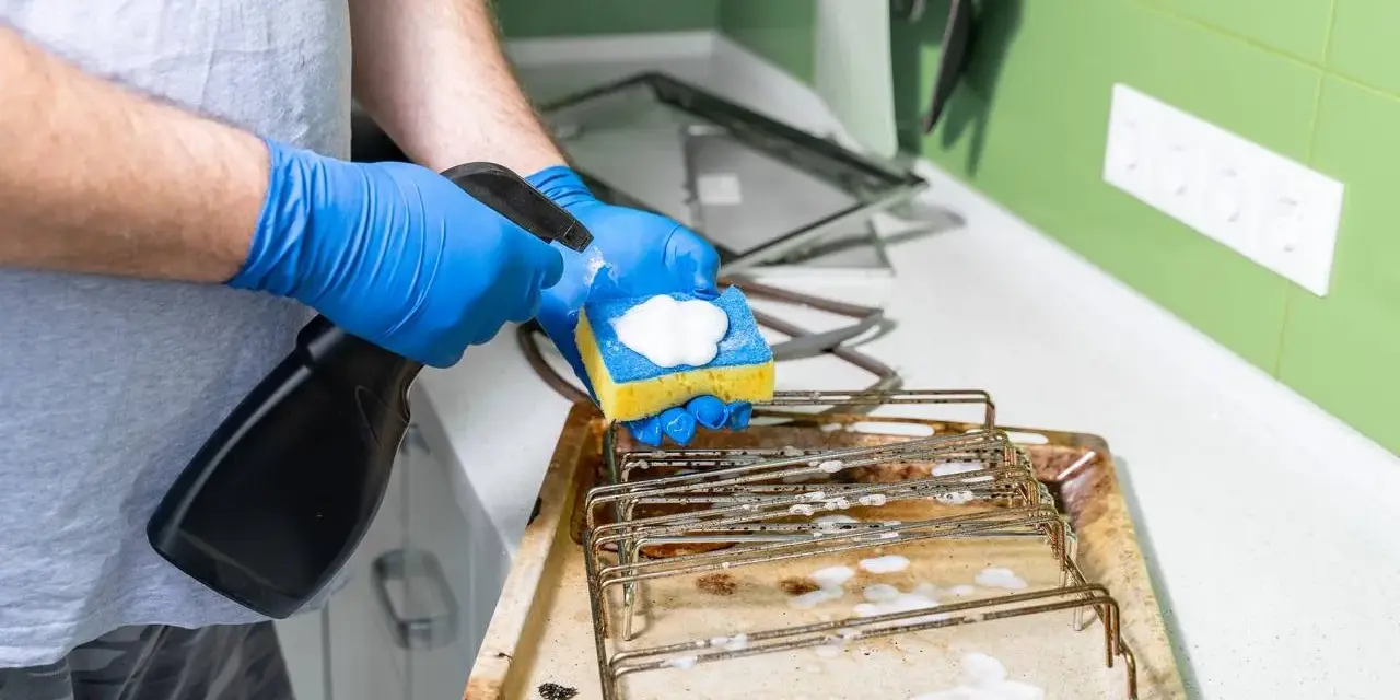 Person mit blauen Handschuhen sprüht Reinigungsmittel auf einen Schwamm, um ein schmutziges Backblech zu reinigen.