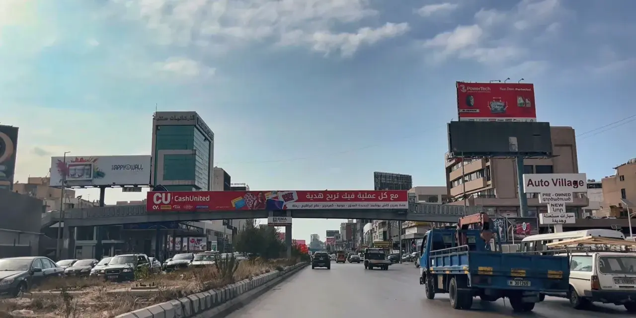 Driving in Lebanon, a busy street with cars, trucks, and billboards advertising various services and products.