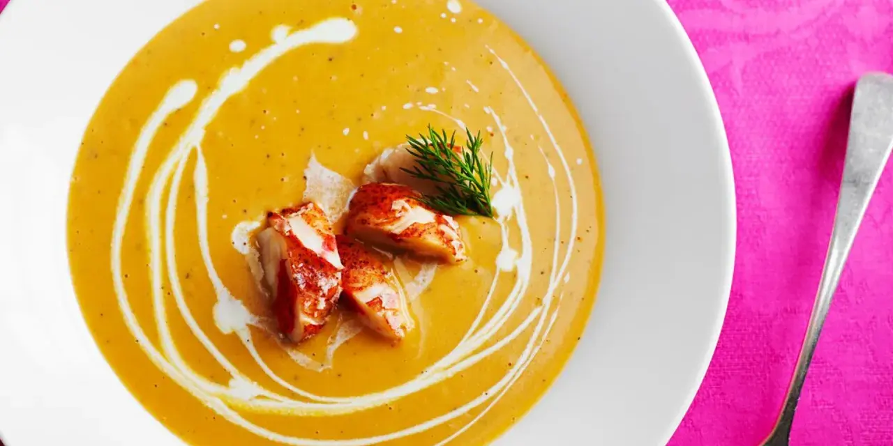 Un cremoso bisque de langosta, adornado con trozos de carne de langosta y un toque de eneldo, servido en un plato blanco sobre un mantel rosa vibrante.