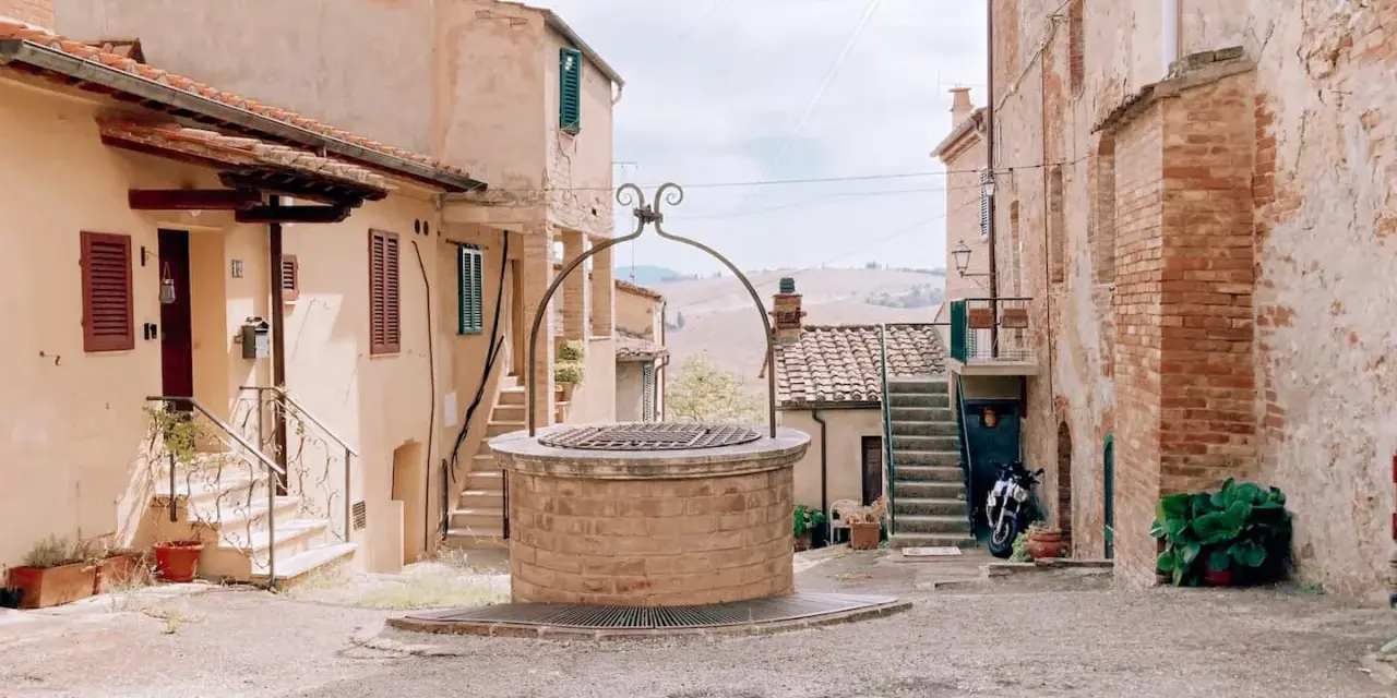 Un antico pozzo in mattoni domina una piazzetta di San Giovanni d'Asso, circondata da case in pietra e con dolci colline sullo sfondo.