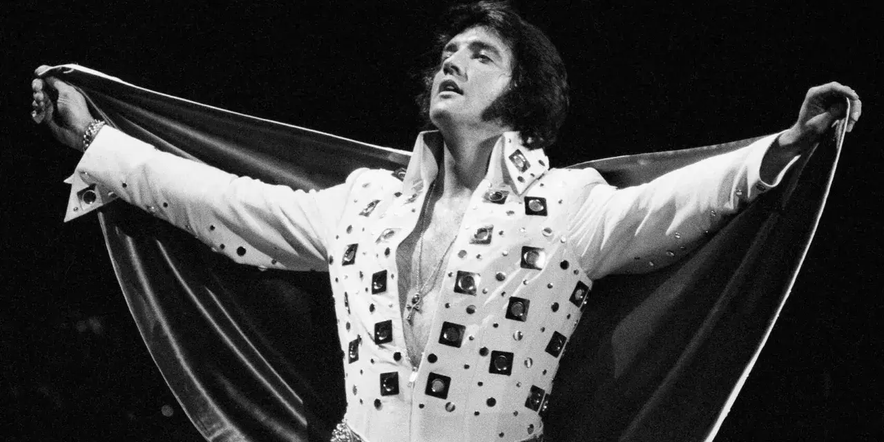 Elvis Presley, der unzählige Tonträger verkaufte, posiert in einem glitzernden weißen Anzug mit ausgebreitetem Umhang.