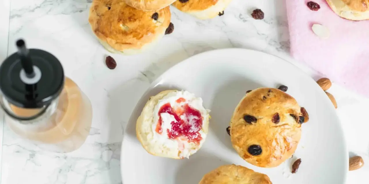 Goldbraune Scones mit Rosinen und Mandeln, ein Scones Teig mit Marmelade und Sahne, daneben ein Getränk.