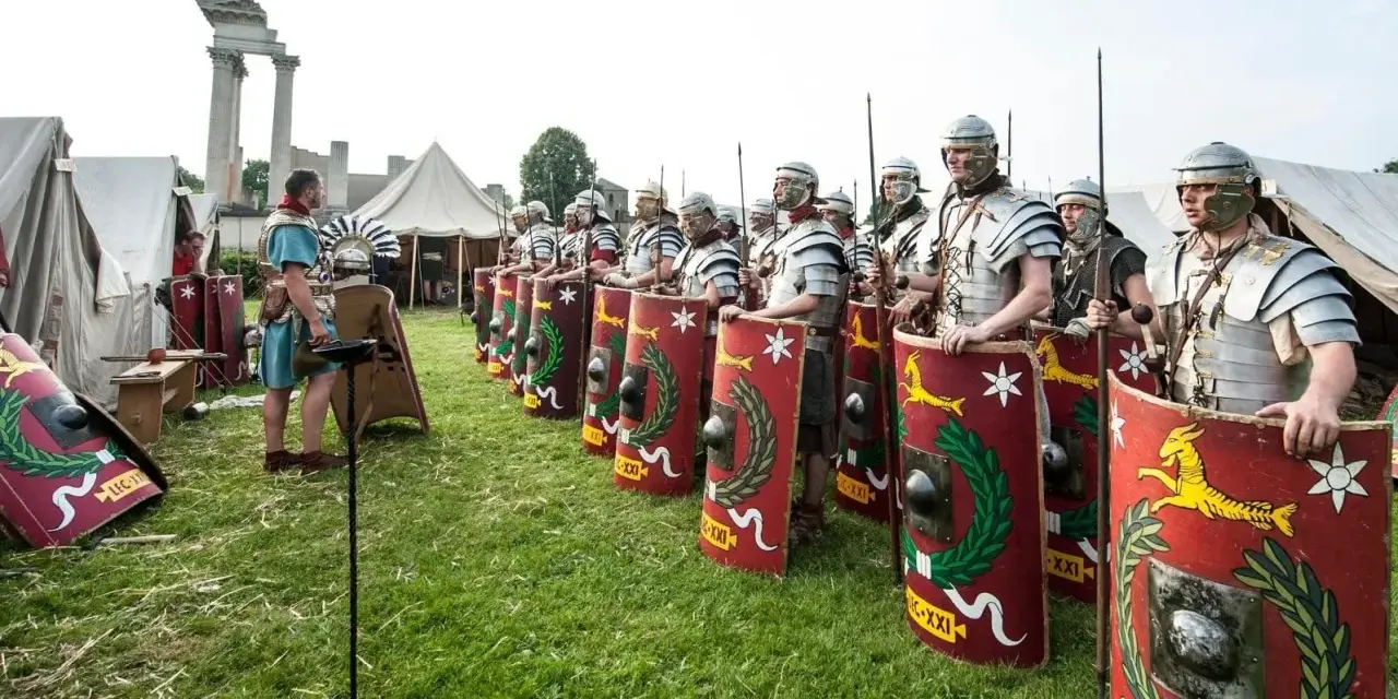 Rzymscy legioniści w pełnym uzbrojeniu, z tarczami i włóczniami, gotowi do bitwy. W tle namioty i ruiny.