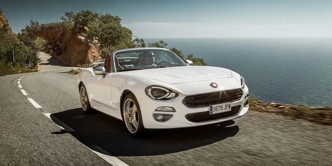 Biały Fiat 124 Spider sunie po krętej drodze nad morzem.