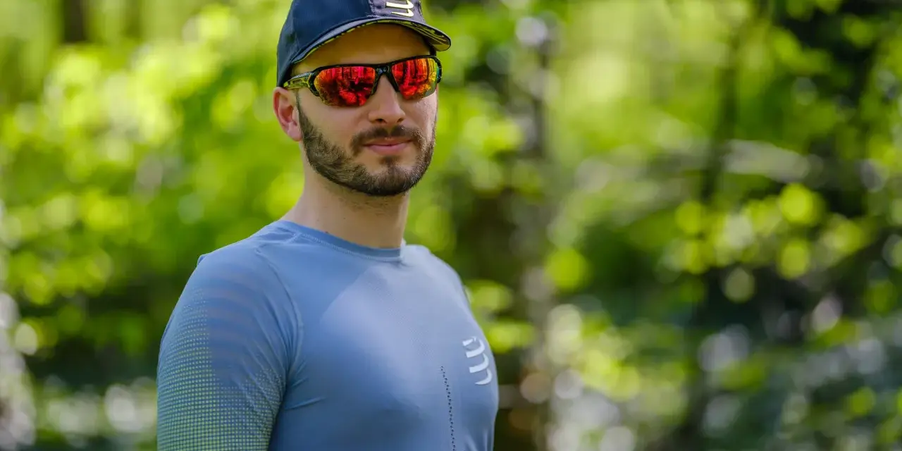 Mężczyzna w czapce i sportowych okularach przeciwsłonecznych, z odbiciem lasu w soczewkach.