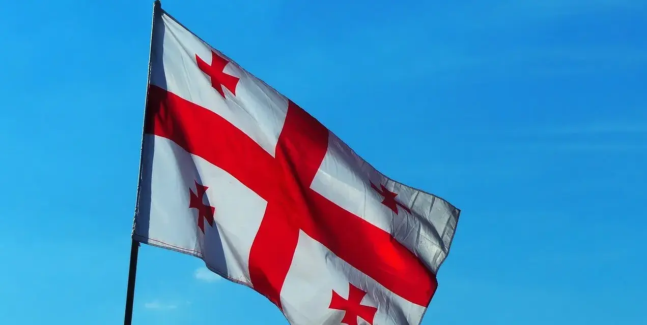 Flaga Gruzji powiewa na tle błękitnego nieba. Białe tło przecina czerwony krzyż, a w każdym z czterech pól znajduje się mniejszy czerwony krzyżyk.