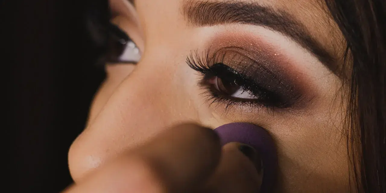 Un œil maquillé avec un smoky eyes facile, rehaussé de paillettes. Une éponge violette est utilisée pour estomper le fond de teint.