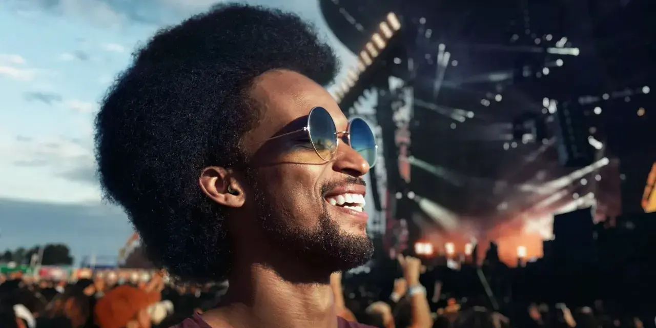 Un homme souriant avec des lunettes de soleil et des bouchons d'oreilles, profitant d'un concert en plein air. Le port des bouchons d'oreilles est obligatoire pour protéger son audition.