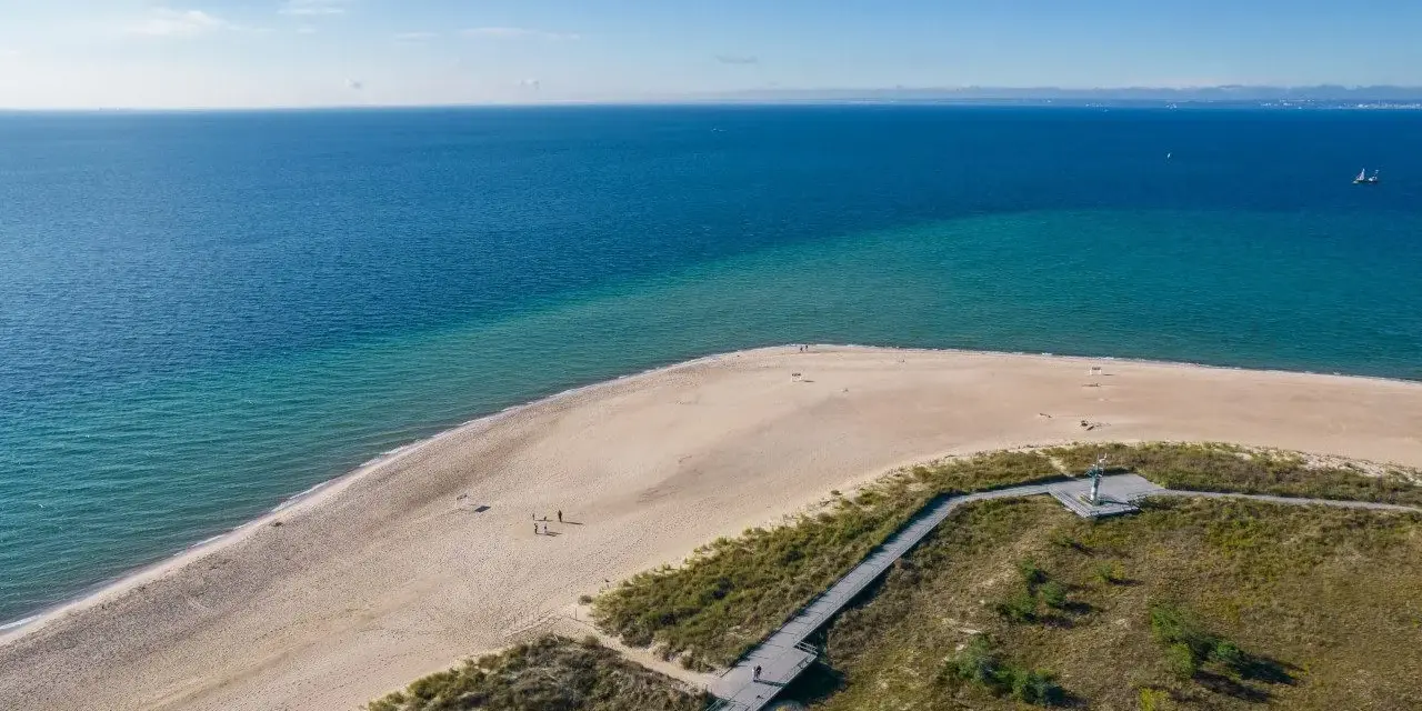 Piękne wydmy helskie nad turkusowym morzem. Drewniana kładka prowadzi przez zarośla na piaszczystą plażę, gdzie spacerują nieliczni turyści.